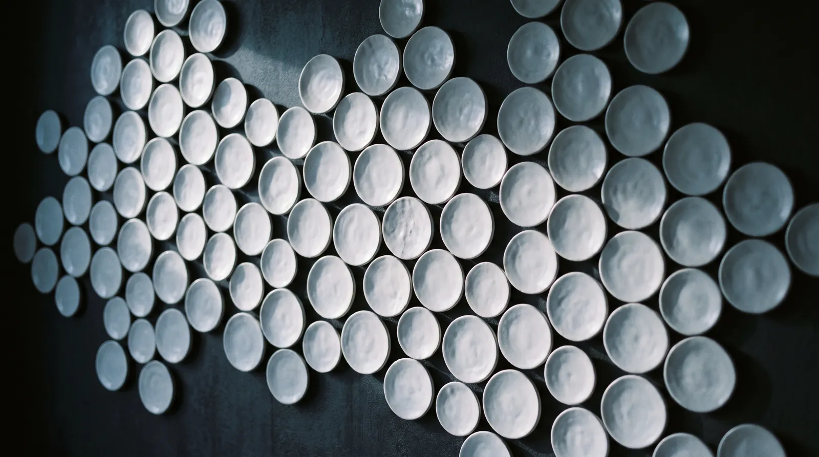 Wall installation of hundreds of handmade white ceramic discs in a honeycomb pattern under raking light