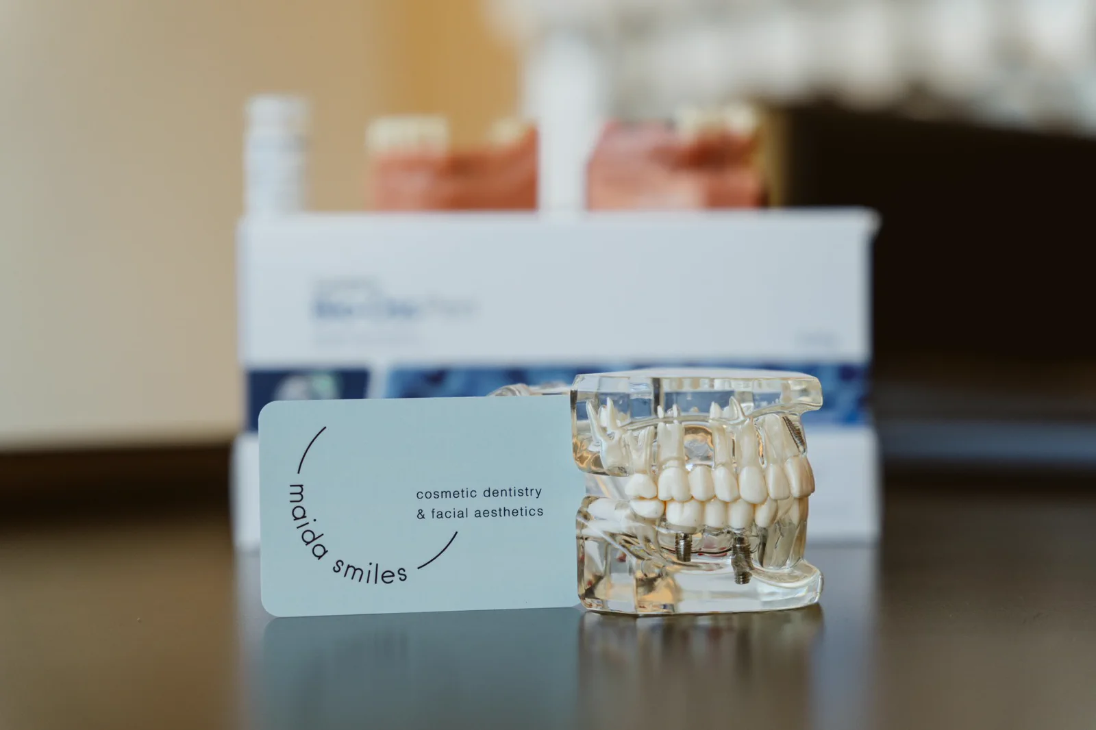 A transparent dental implant model stands beside a Maida Smiles card.