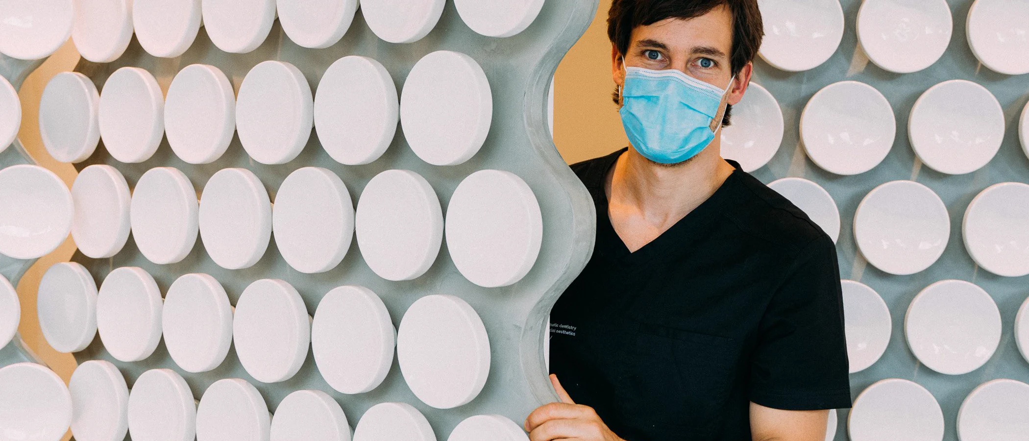 A masked dentist stands beside the ceramic feature wall at Maida Smiles.