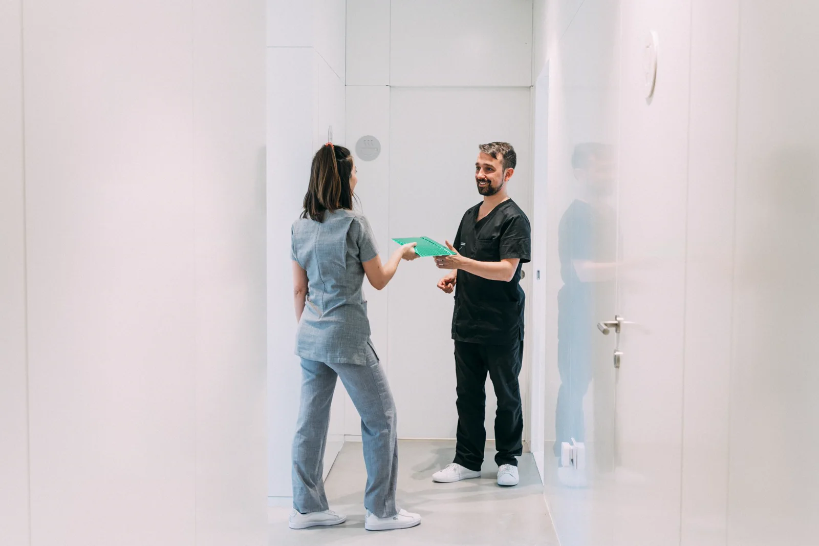 Two colleagues exchange a patient folder in a bright clinic corridor.
